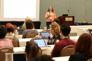 A female speaker is standing at the front of a room speaking to 5 rows of attendees, who are sitting at long white tables and taking notes on laptops, tablets and on paper.