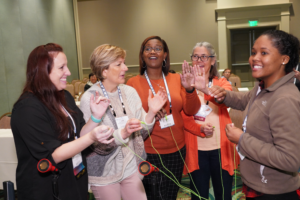 Four women are smiling while testing a technologies that has wires connecting each of their palms. a fifth woman is standing to their right and smiling at the camera.
