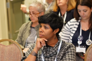 A female attendee is sitting in an education session and looking attentively towards the front of the room, with two female attendees sitting in the row behind her and taking notes.