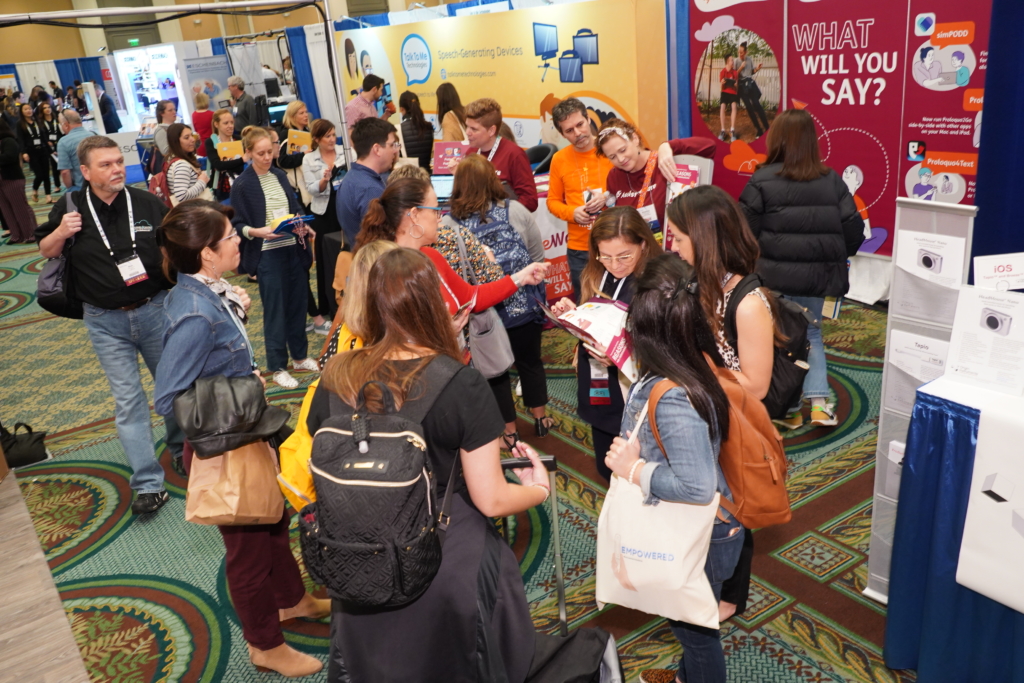 A large group of more than 20 people is gathered in the ATIA exhibit hall looking at exhibiting booths. 