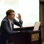 Speaker with glasses stands in front of podium and gestures while speaking in front of audience.
