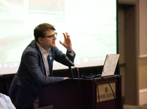 Speaker with glasses stands in front of podium and gestures while speaking in front of audience.