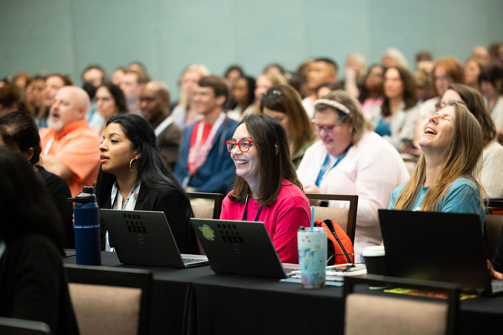A large crowd of attendees at ATIA 2023 listens to a speaker. A woman in hot pink glasses smiles while sitting in front of a laptop.
