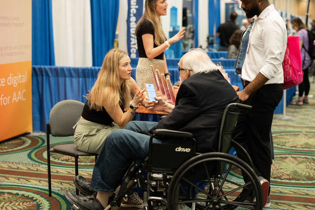 An exhibitor at ATIA 2023 is sitting in a chair and leans forward to speak to an attendee in a wheelchair.