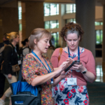 Two ATIA female attendees look at their mobile phones as they access the latest conference updates on the ATIA mobile app.