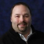 Headshot of Brian Wojcik wearing a black zip up sweater with a white button up shirt underneath it.