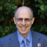 Picture of Neil Charness wearing a blue button up shirt with a blue tie over a dark blue sports coat.